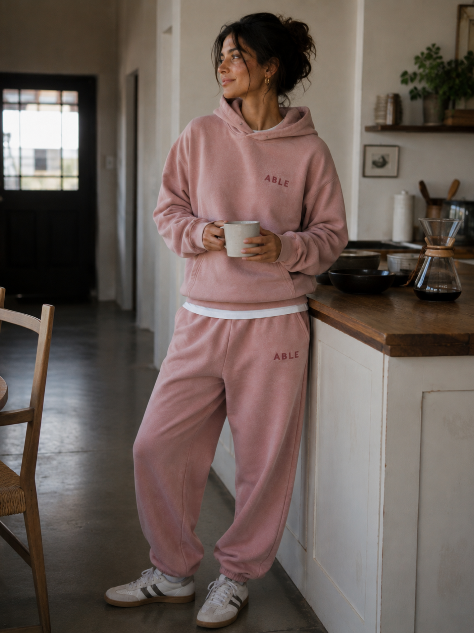 A woman wearing a matching dusty pink loungewear set with the word 'ABLE' embroidered on the chest and thigh, holding a white mug in a kitchen setting.