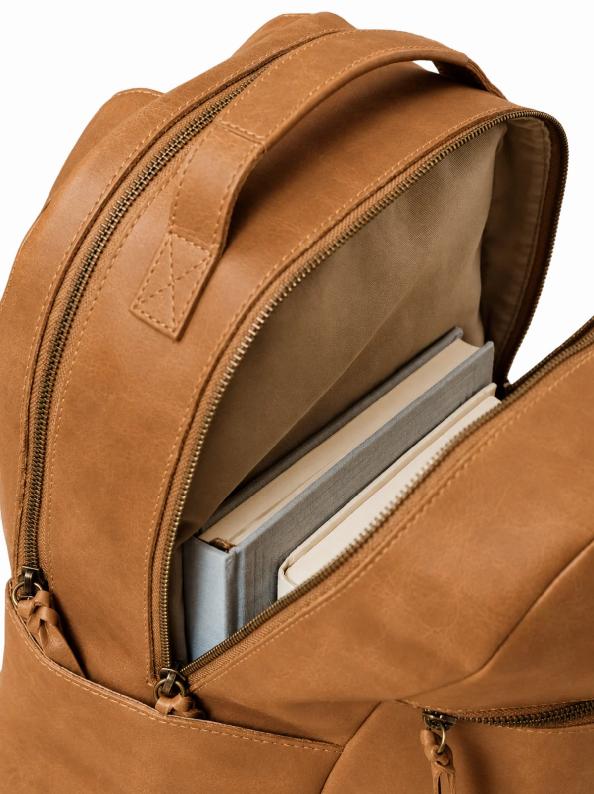 Close-up of a tan leather backpack with its top front zipper open, showing books and notebooks inside against a white background. #Color_Camel