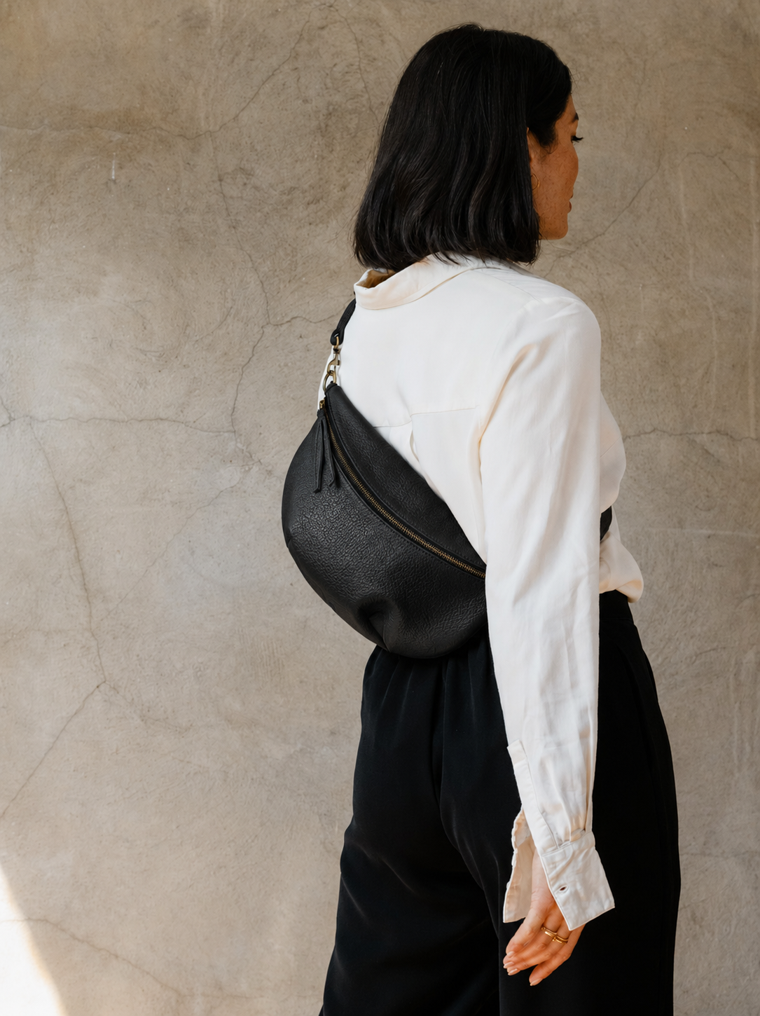 Woman wearing a pebbled textured leather black belt bag over one shoulder across their back against a beige wall #Color_Pebbled Black