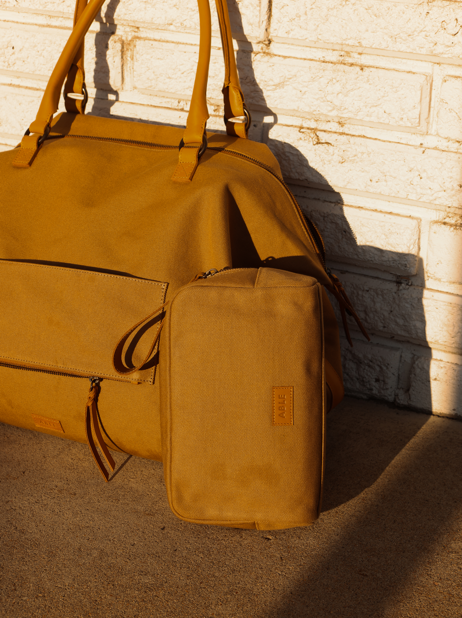 Tan canvas weekender bag leaning against a textured wall with tan canvas dopp kit against it.