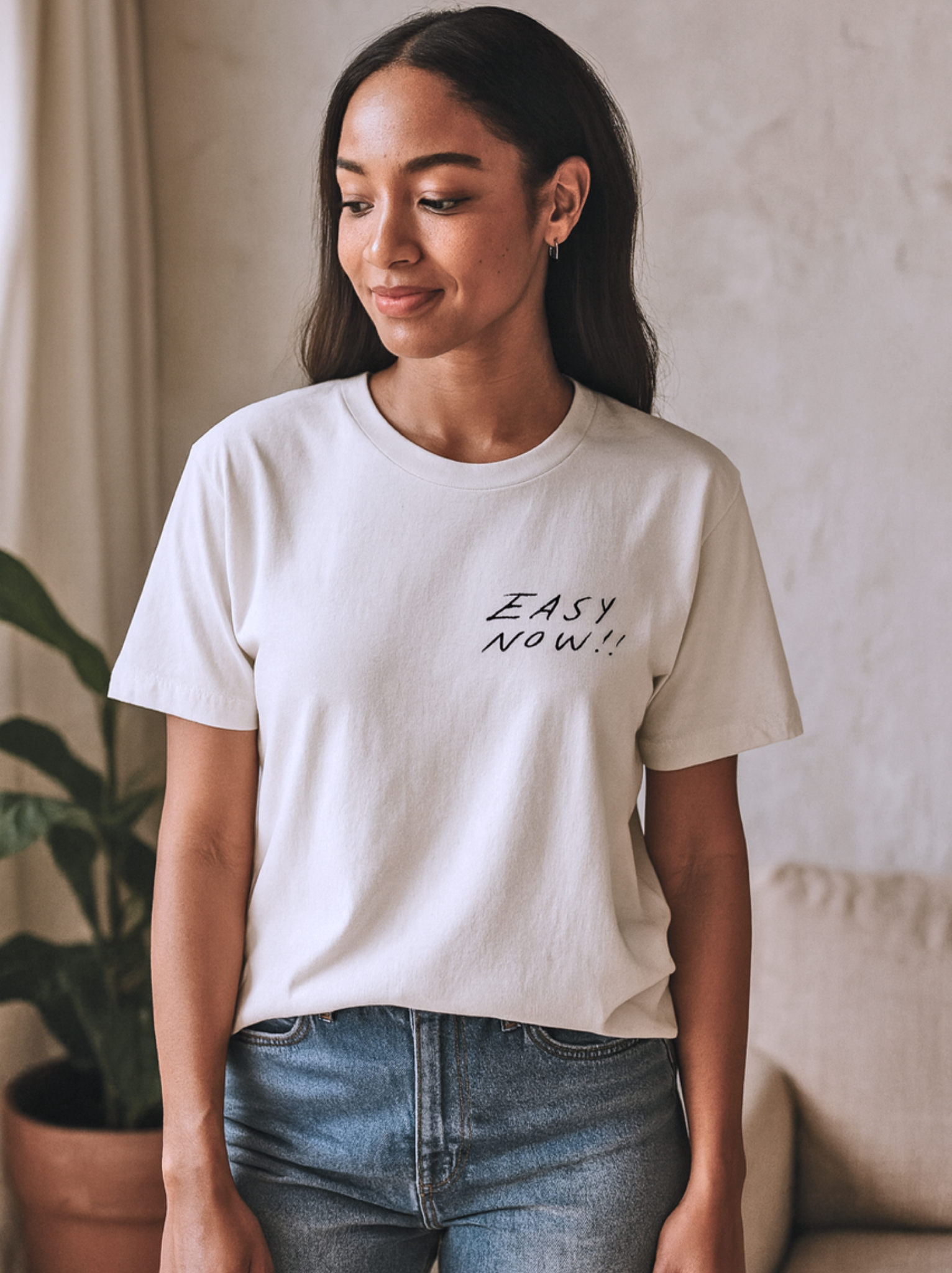 Woman wearing a Easy Now Tee
white t-shirt with 'EASY NOW!' text, standing indoors.