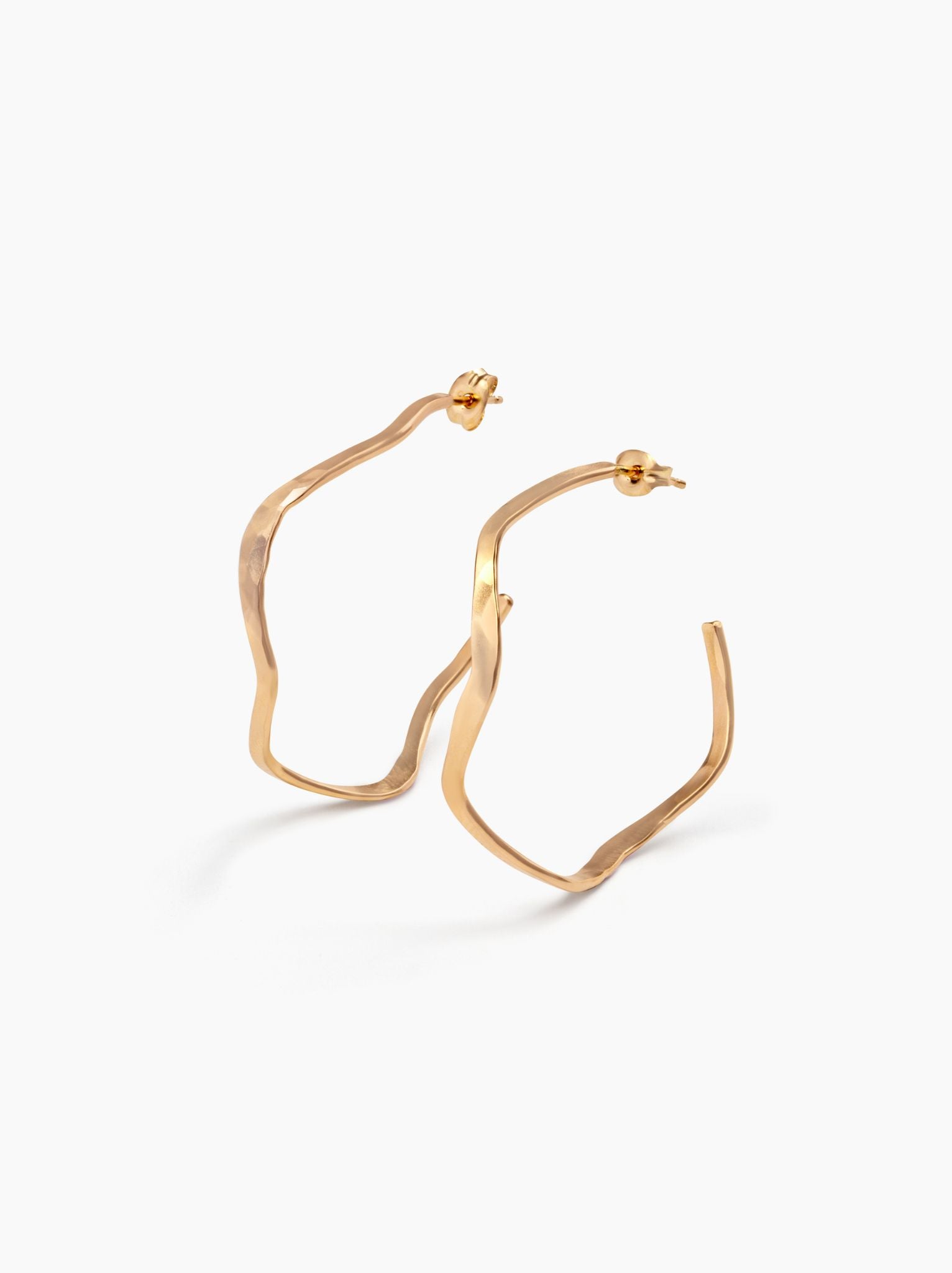 Side angle of gold wavy hoop earrings with ear posts on a white background.