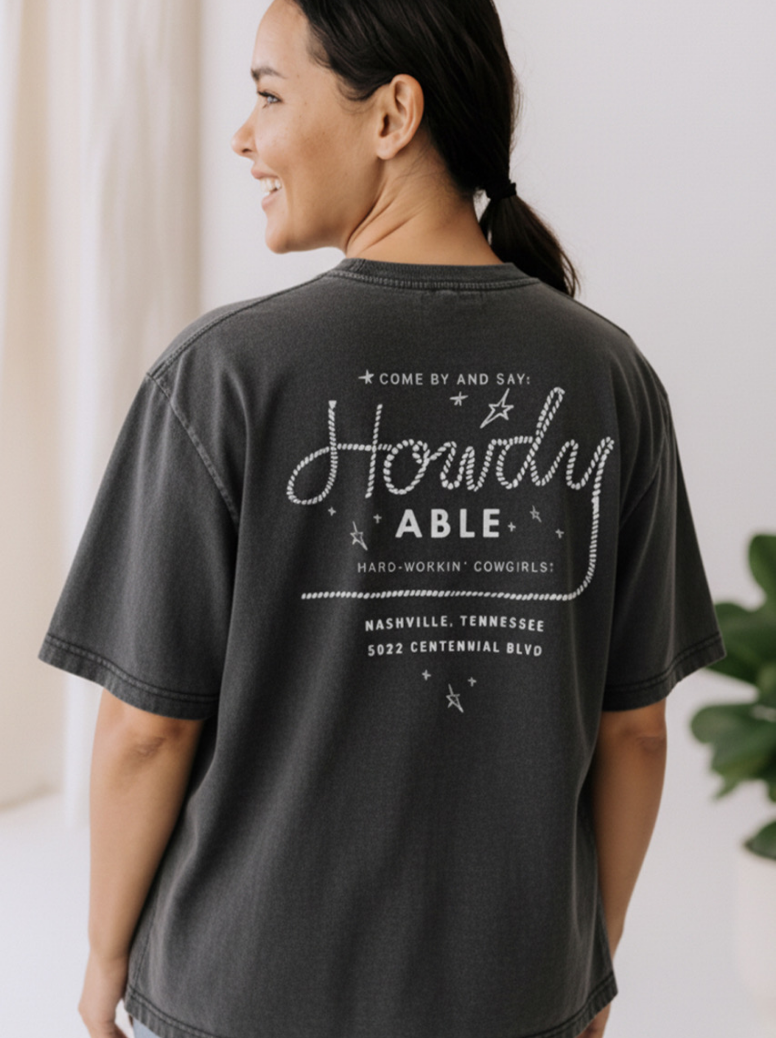 Person wearing a black Say Hi Tee t-shirt with text on the back, standing indoors.