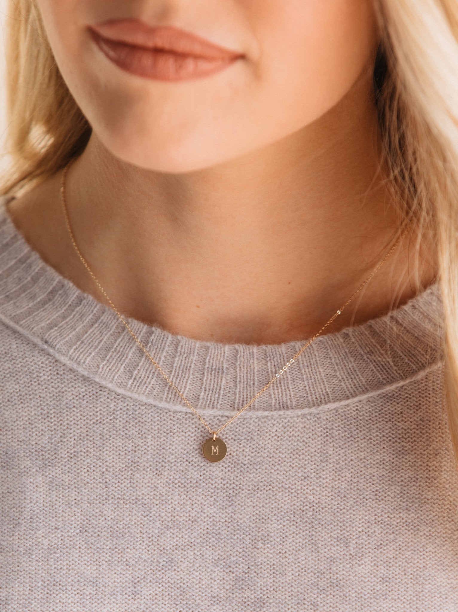 Close up of woman a wearing gold necklace with small round pendant featuring the letter 'M' and a sweater.