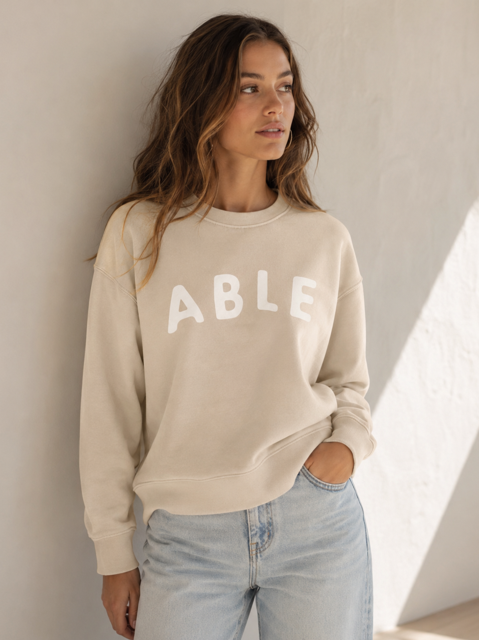 A woman wearing a cream-colored crewneck sweatshirt with the word 'ABLE' printed in white across the chest, paired with light-wash blue jeans, standing against a textured off-white wall.