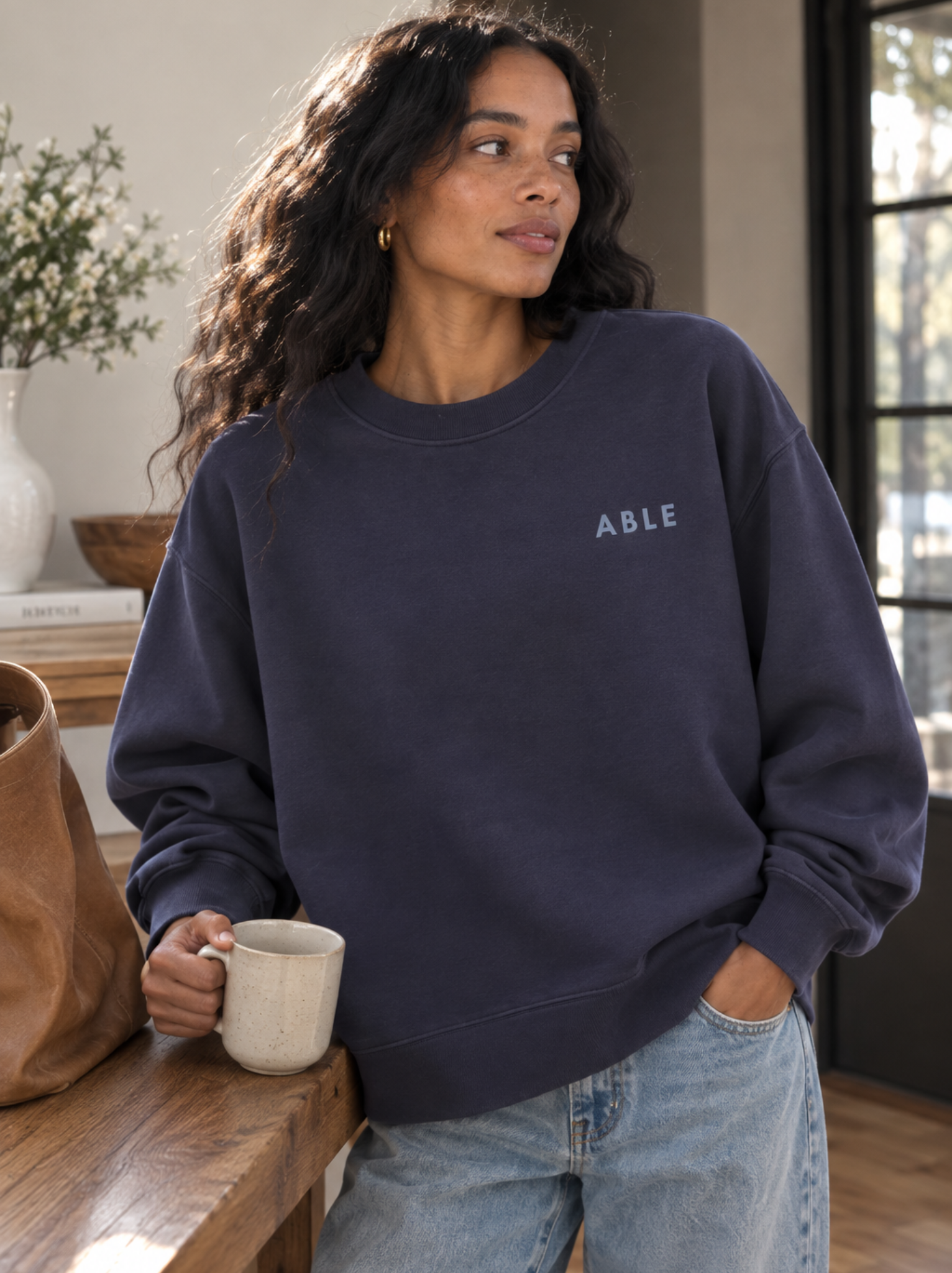 A woman wearing a navy blue crewneck sweatshirt with the word 'ABLE' printed in light blue on the left chest, paired with light-wash blue jeans, holding a ceramic mug in a warm indoor setting. #Color_Navy Ink