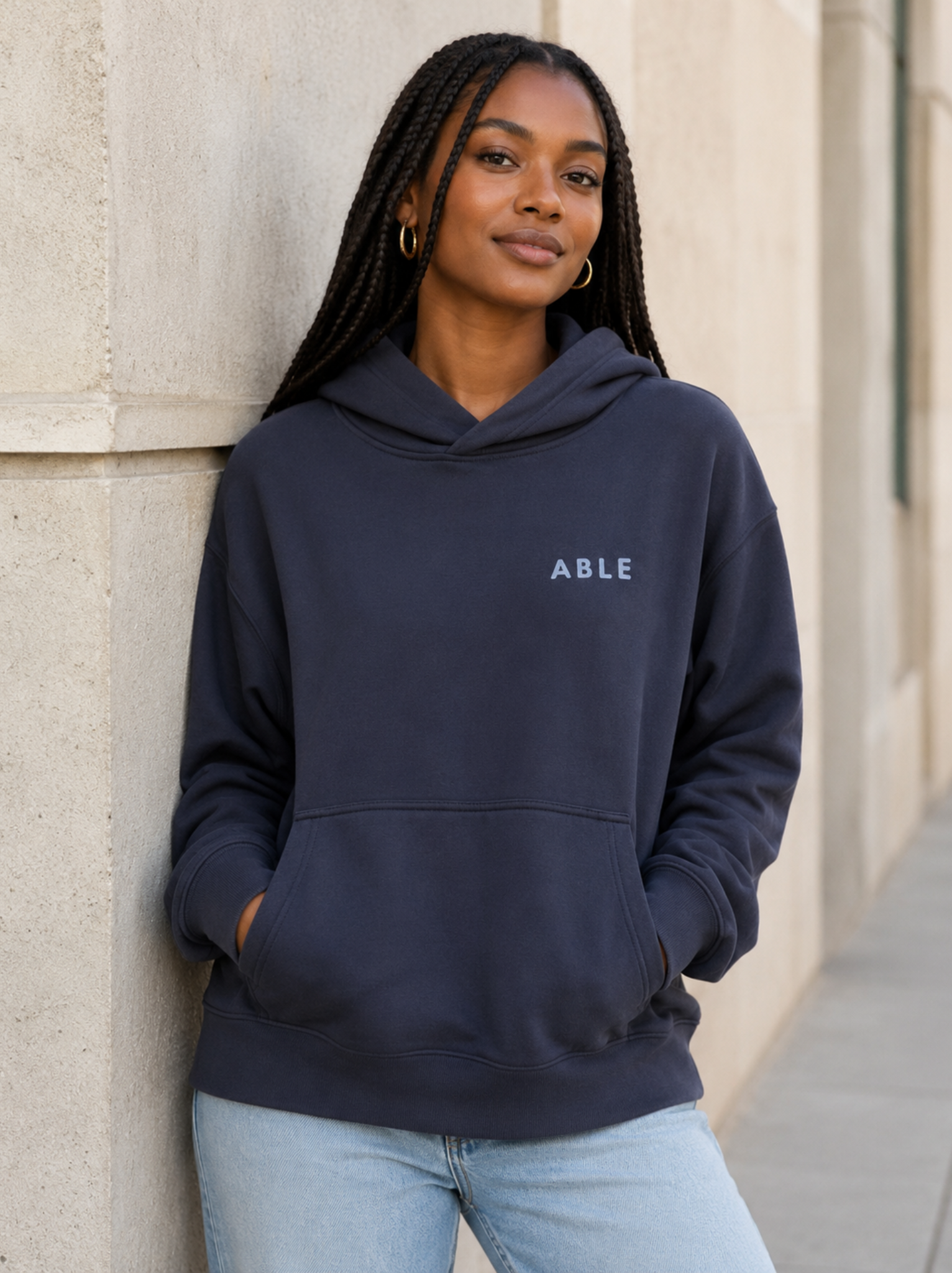 A woman with long black braids leans against a light beige stone wall, wearing a navy blue pullover hoodie with the word 'ABLE' printed in white on the left chest, paired with light blue jeans. #Color_Navy Ink