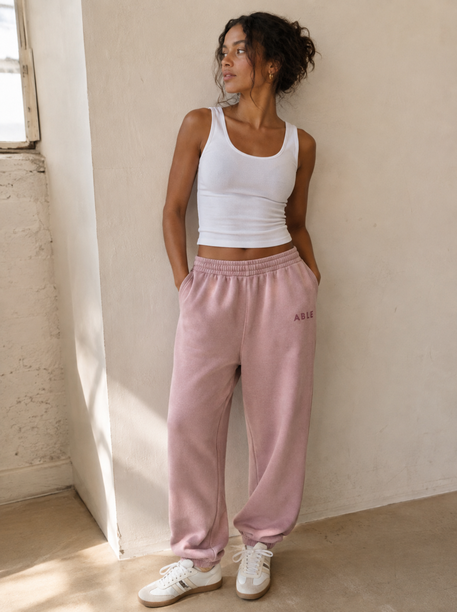 A woman stands in a sunlit room wearing a white ribbed tank top and dusty pink sweatpants with 'ABLE' printed on the thigh, paired with white sneakers.