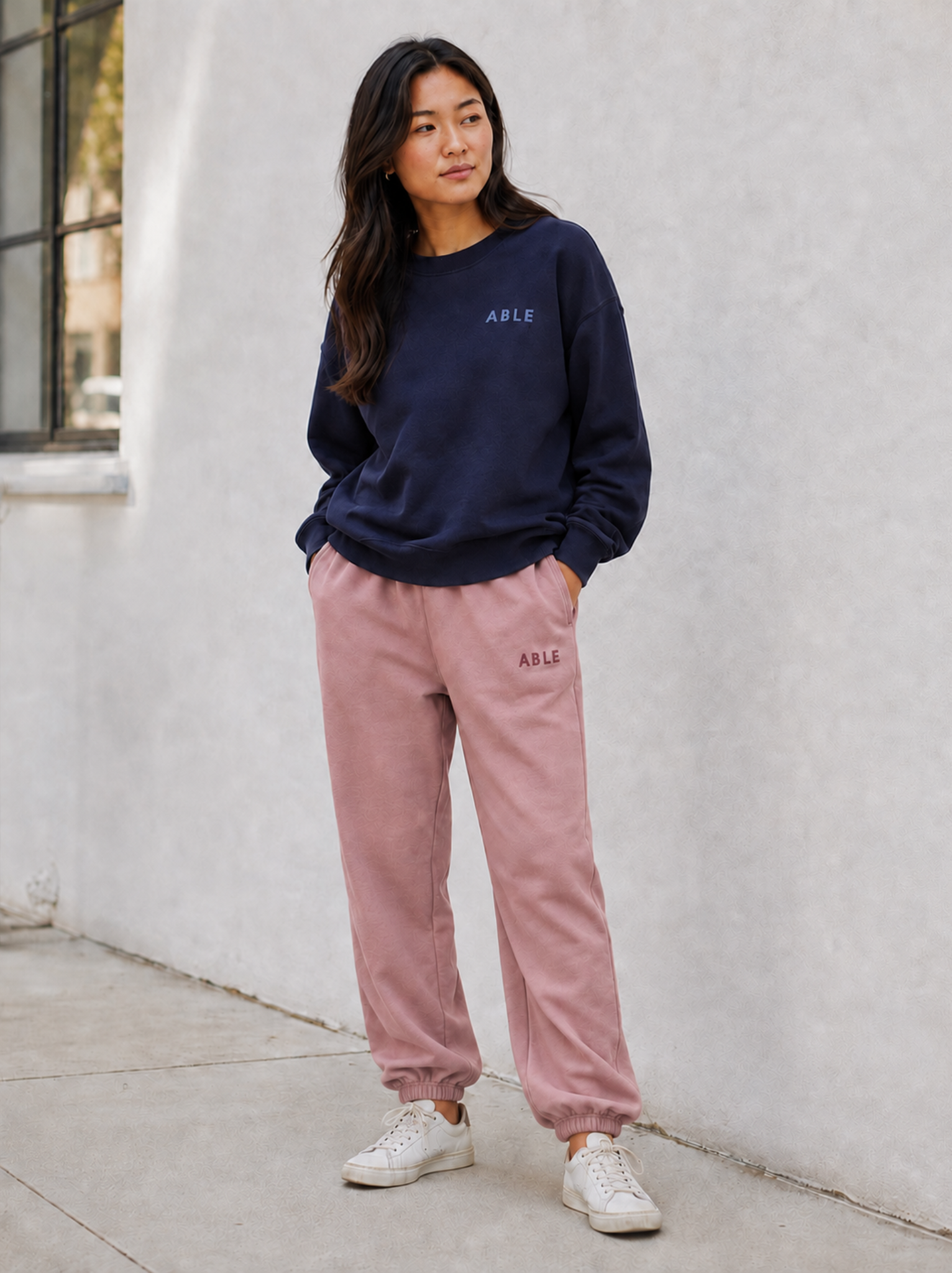 A woman stands outdoors against a light stucco wall wearing a navy crewneck sweatshirt and dusty pink jogger pants, both with small 'ABLE' logo on the chest and upper thigh. She is wearing white sneakers.