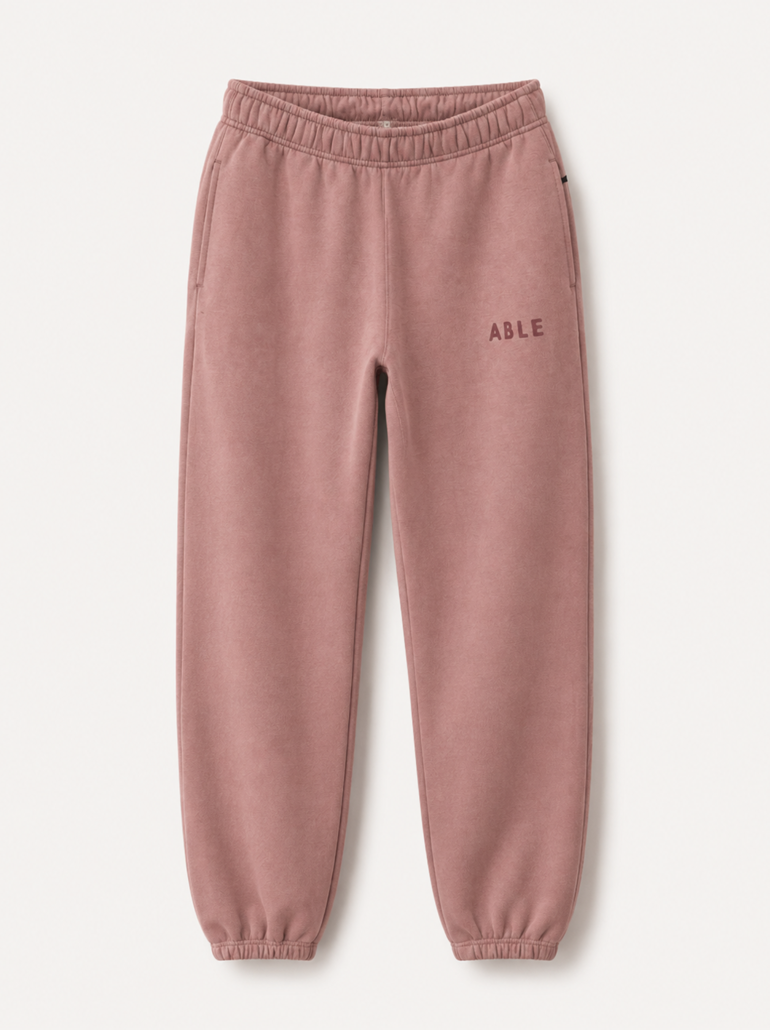 A pair of dusty pink fleece jogger pants with an elastic waistband and cuffs, featuring the word 'ABLE' printed in dark pink on the left hip, shown against a light off-white background.