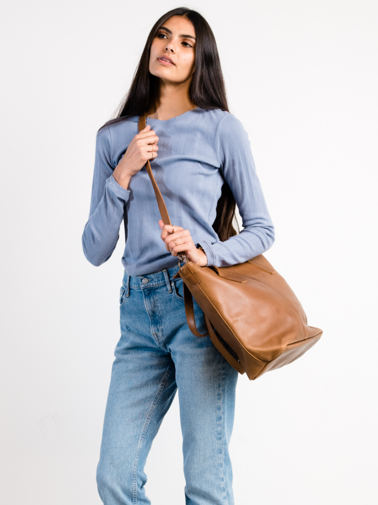 Woman holding a brown leather bag across her body with crossbody strap against a white background #Color_Whiskey