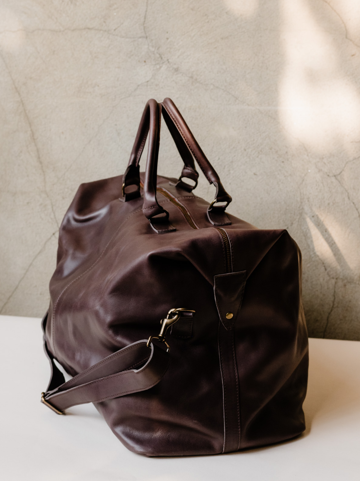 Side view of chocolate brown leather weekender bag, shows side snaps, handles and crossbody strap, sitting on a neutral surface against a grey wall. #Color_Chocolate Brown