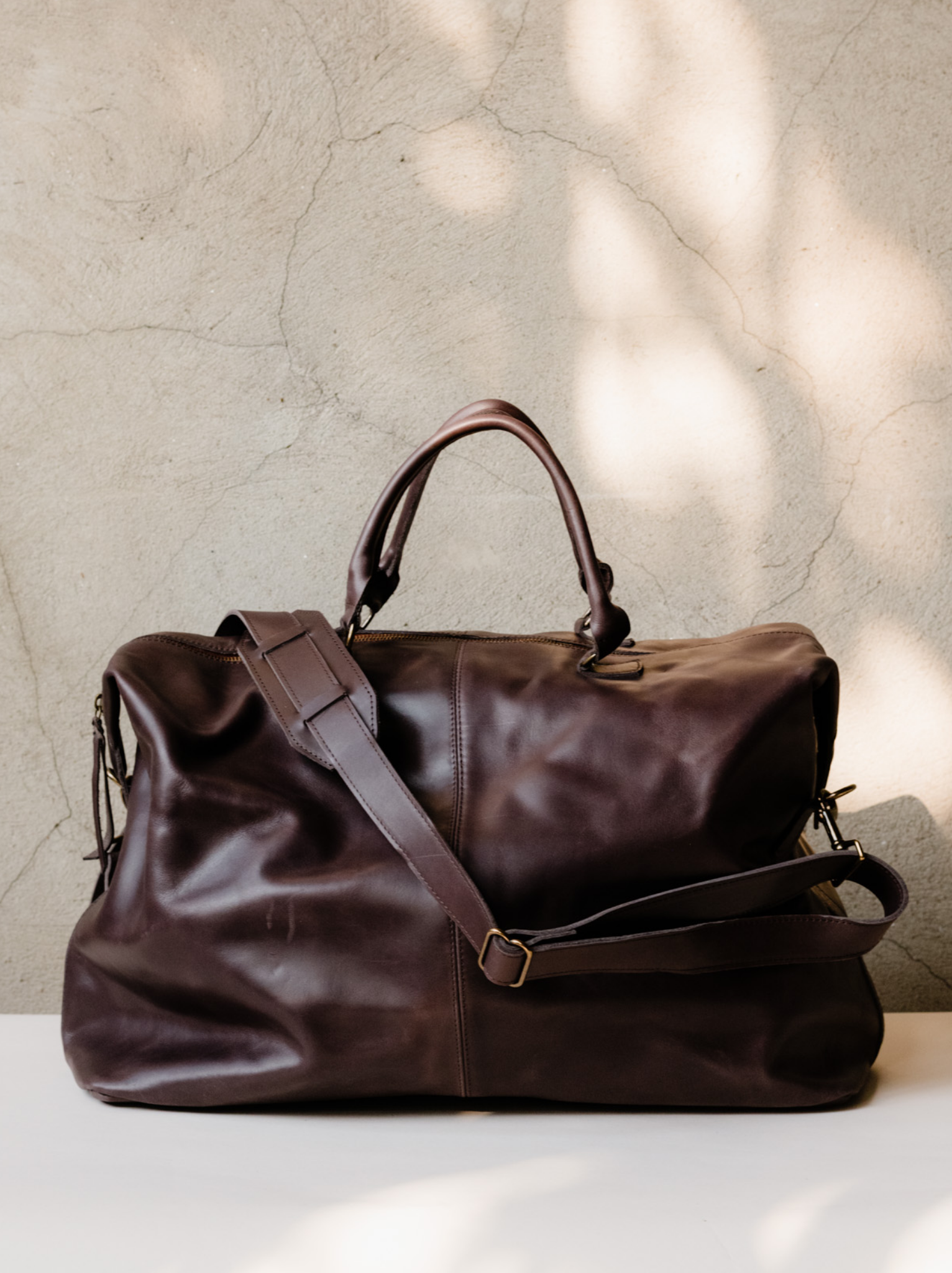 A chocolate brown leather weekender duffel bag sitting on a beige surface against a grey wall. #Color_Chocolate Brown