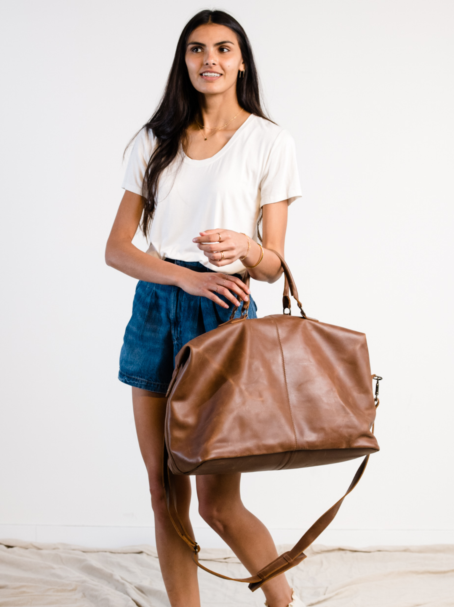 Woman holding a brown leather weekender bag on her arm against a white background. #Color_Whiskey