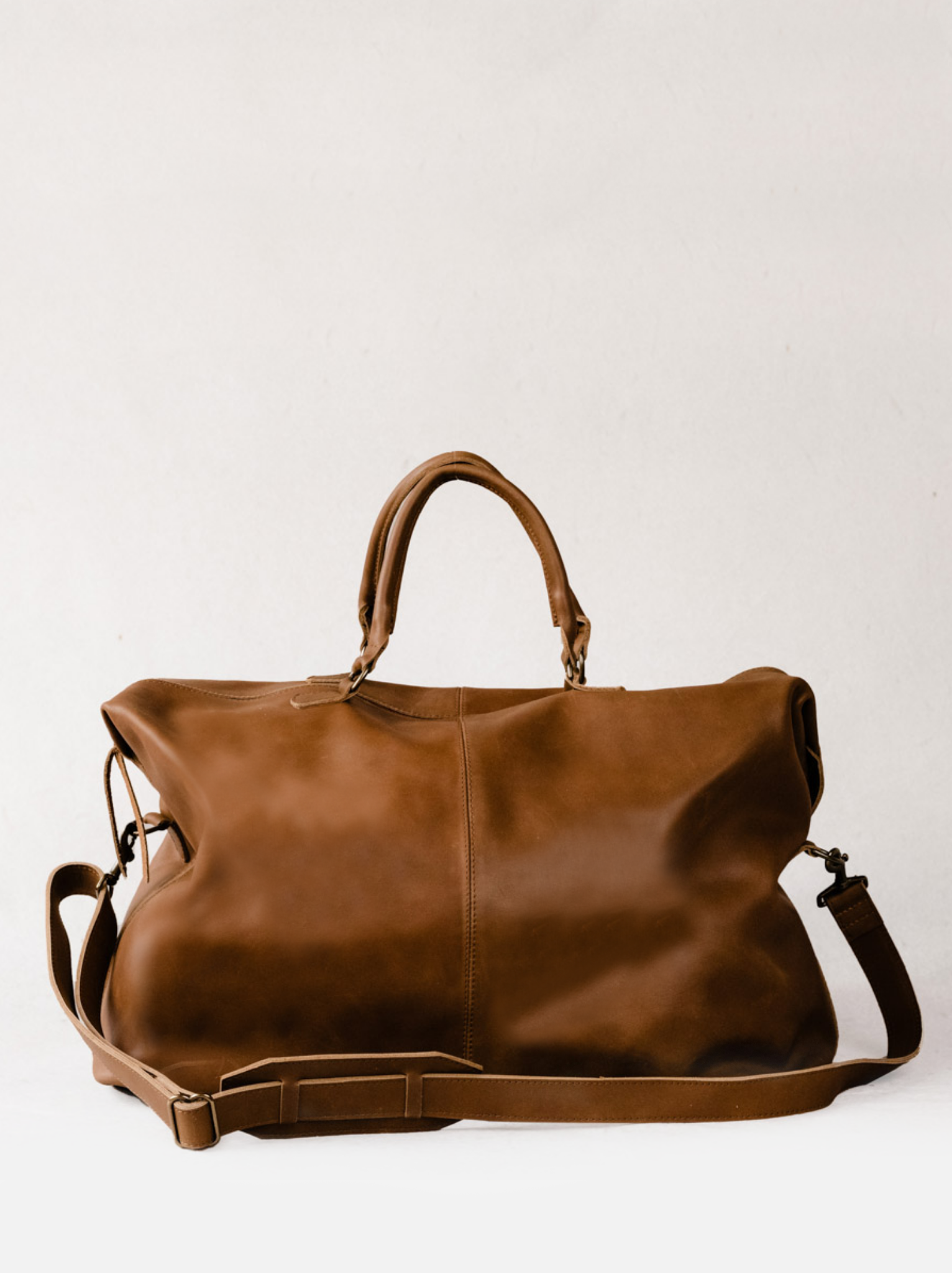 A brown leather weekender duffel bag with side snap closures and a double handle strap and detachable crossbody strap, placed on a white background. #Color_Whiskey
