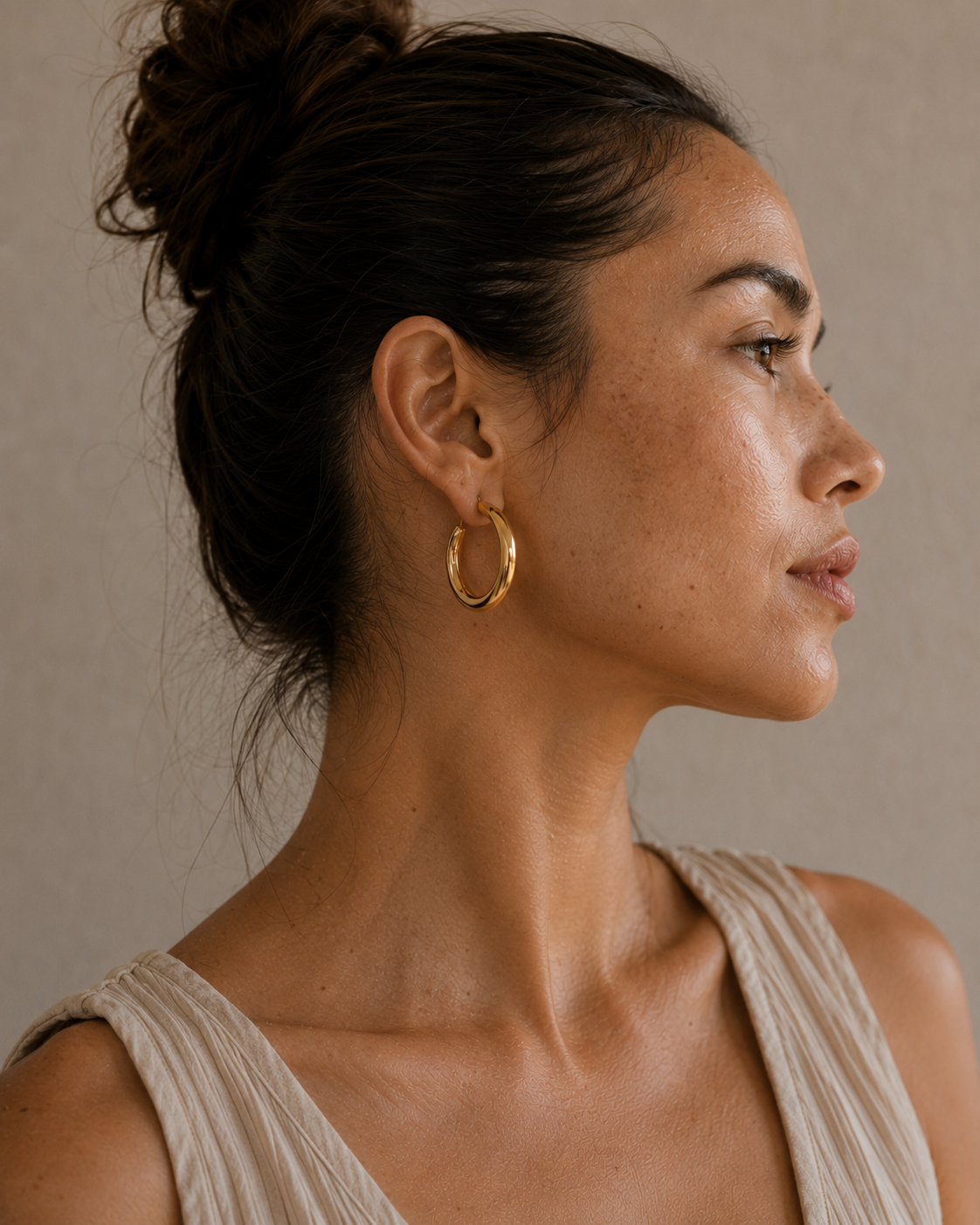 A woman in profile wearing a chunky gold hoop earring, shown against a neutral beige background.