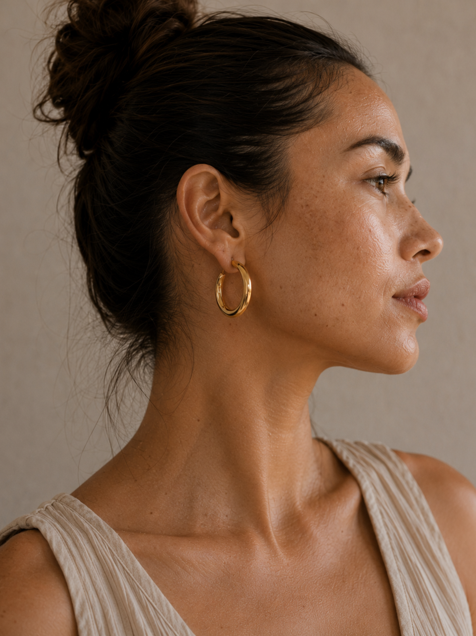 A woman in profile wearing a chunky gold hoop earring, shown against a neutral beige background.