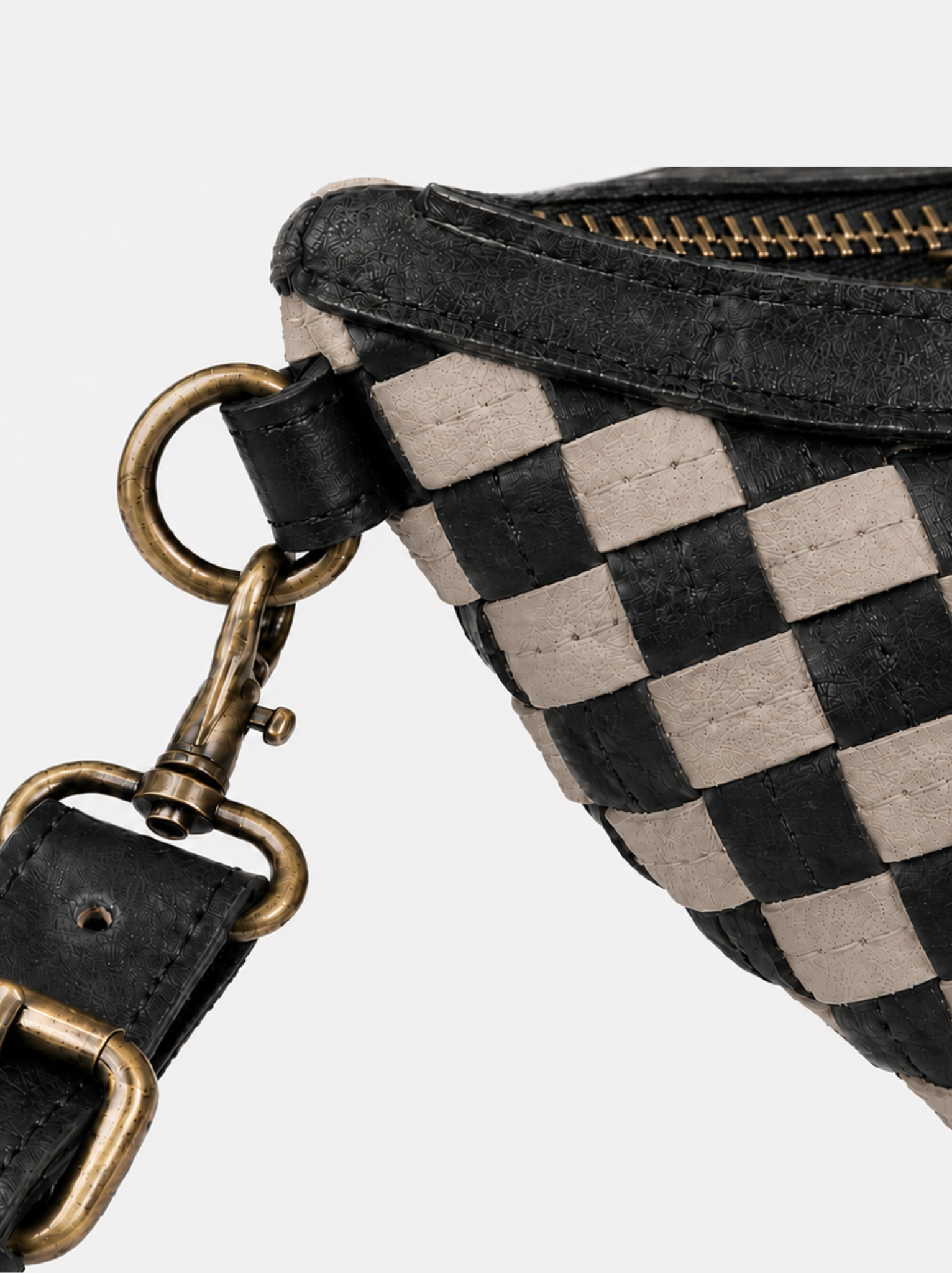Close-up of a woven black and beige checkered bag with a black leather strap and antique bronze-tone metal hardware, set against a plain light gray background.
