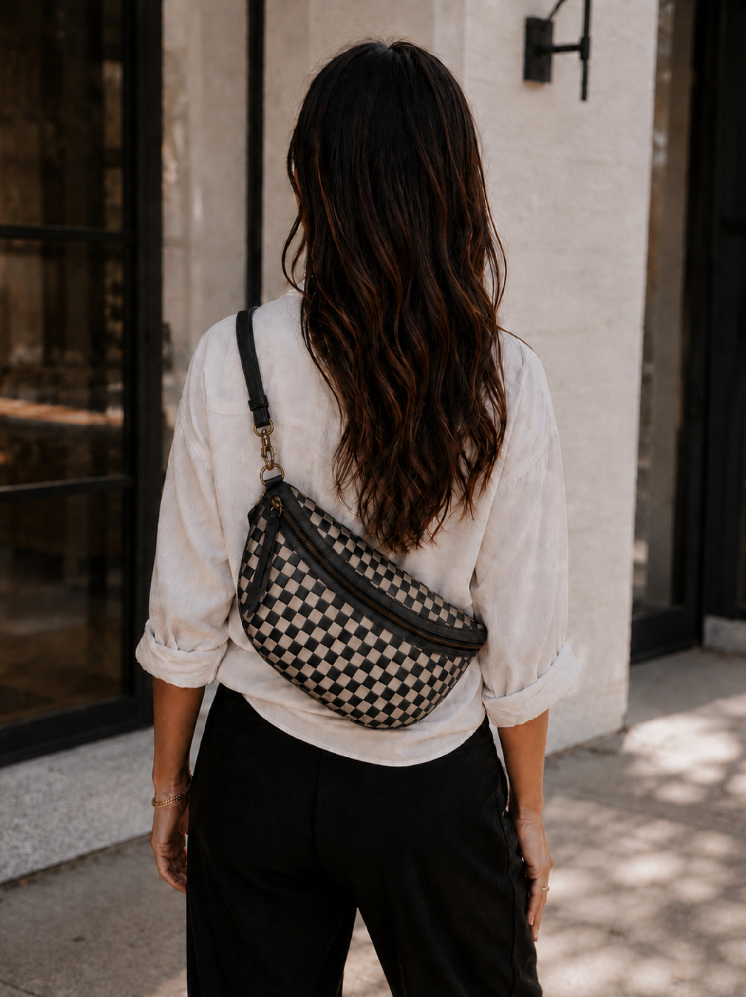 A woman viewed from behind wearing a black and beige checkered belt bag with a black strap, styled with a white blouse and black pants.