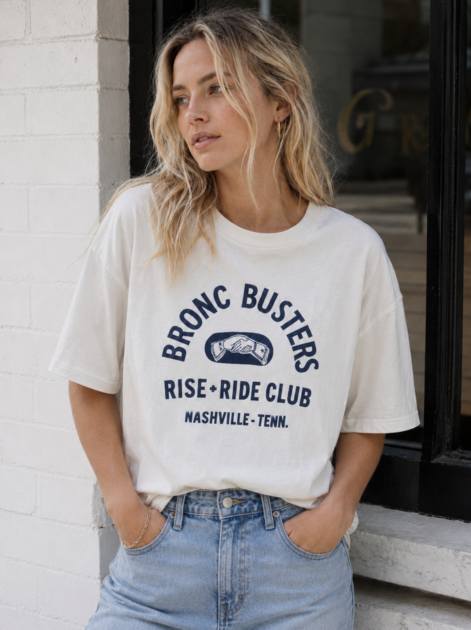 A woman wearing a cream-colored graphic t-shirt with a navy blue 'Bronc Busters Rise-Ride Club Nashville-Tenn.' print, paired with light-wash blue jeans, standing against a white brick wall.