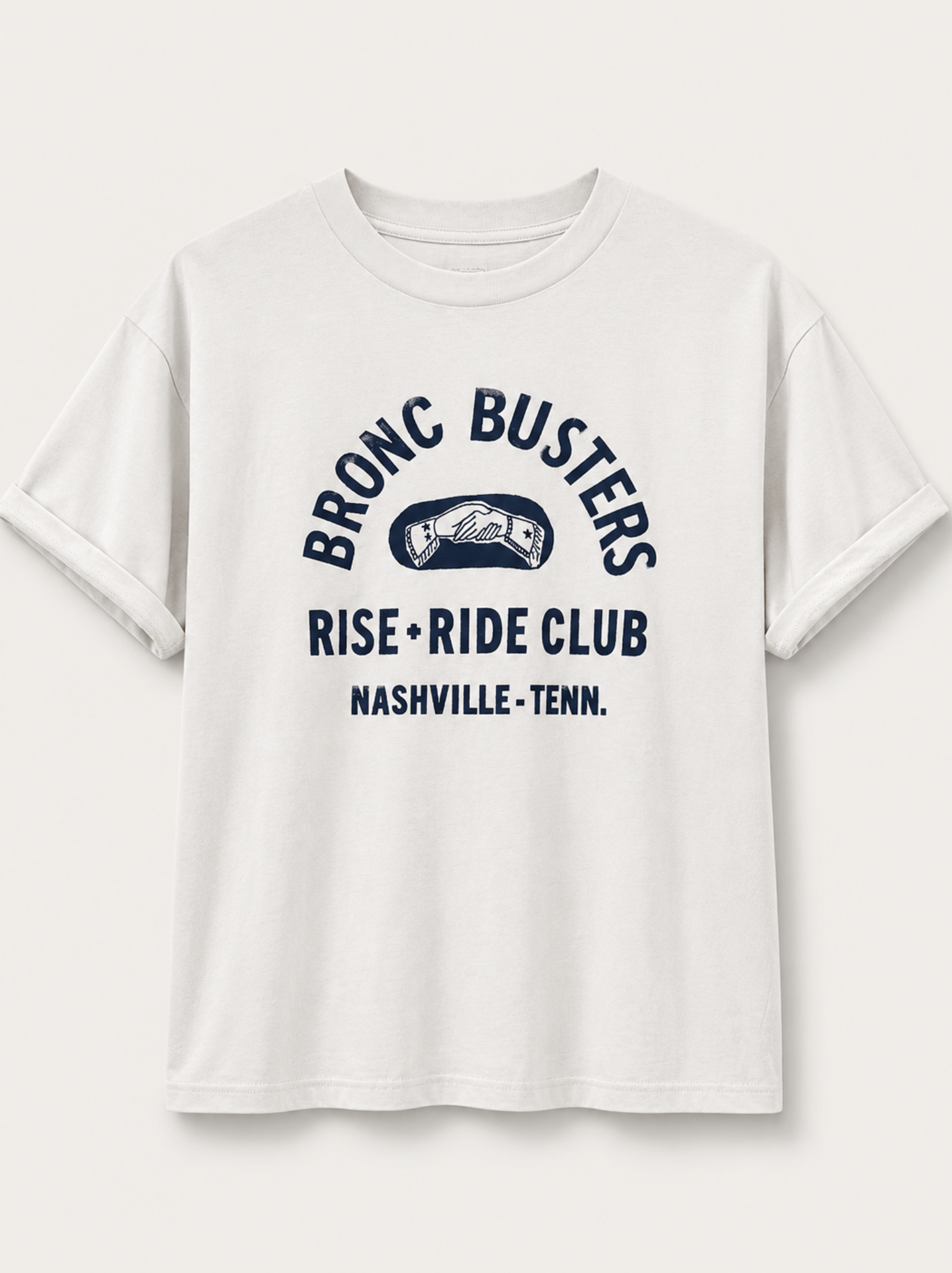 A white short-sleeve crewneck t-shirt with a navy blue graphic print on the front that reads 'BRONC BUSTERS RISE • RIDE CLUB NASHVILLE • TENN.' with a small handshake illustration, shown against a light off-white background.