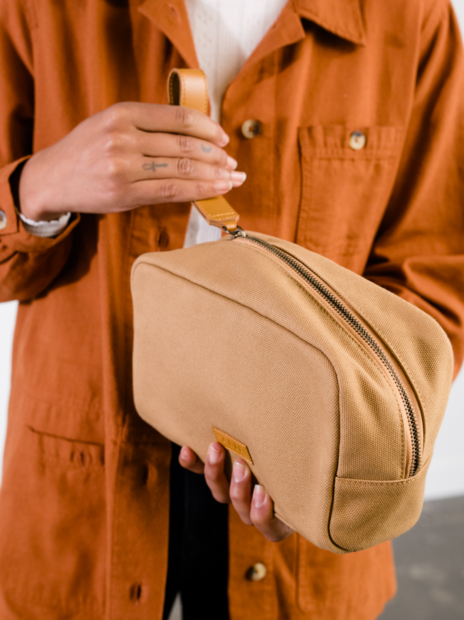 Person unzipping a tan canvas toiletry bag using the cognac leather strap with an orange jacket