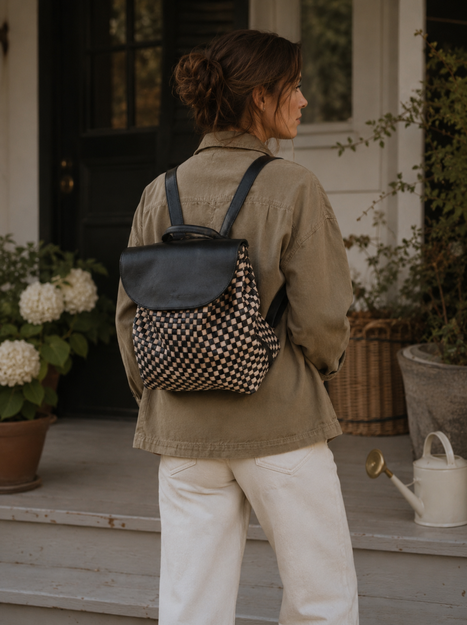 A woman viewed from behind wearing a black and beige checkered backpack with a black leather flap, standing on a porch with potted plants and a white watering can in the background. #Color_Black/Tan