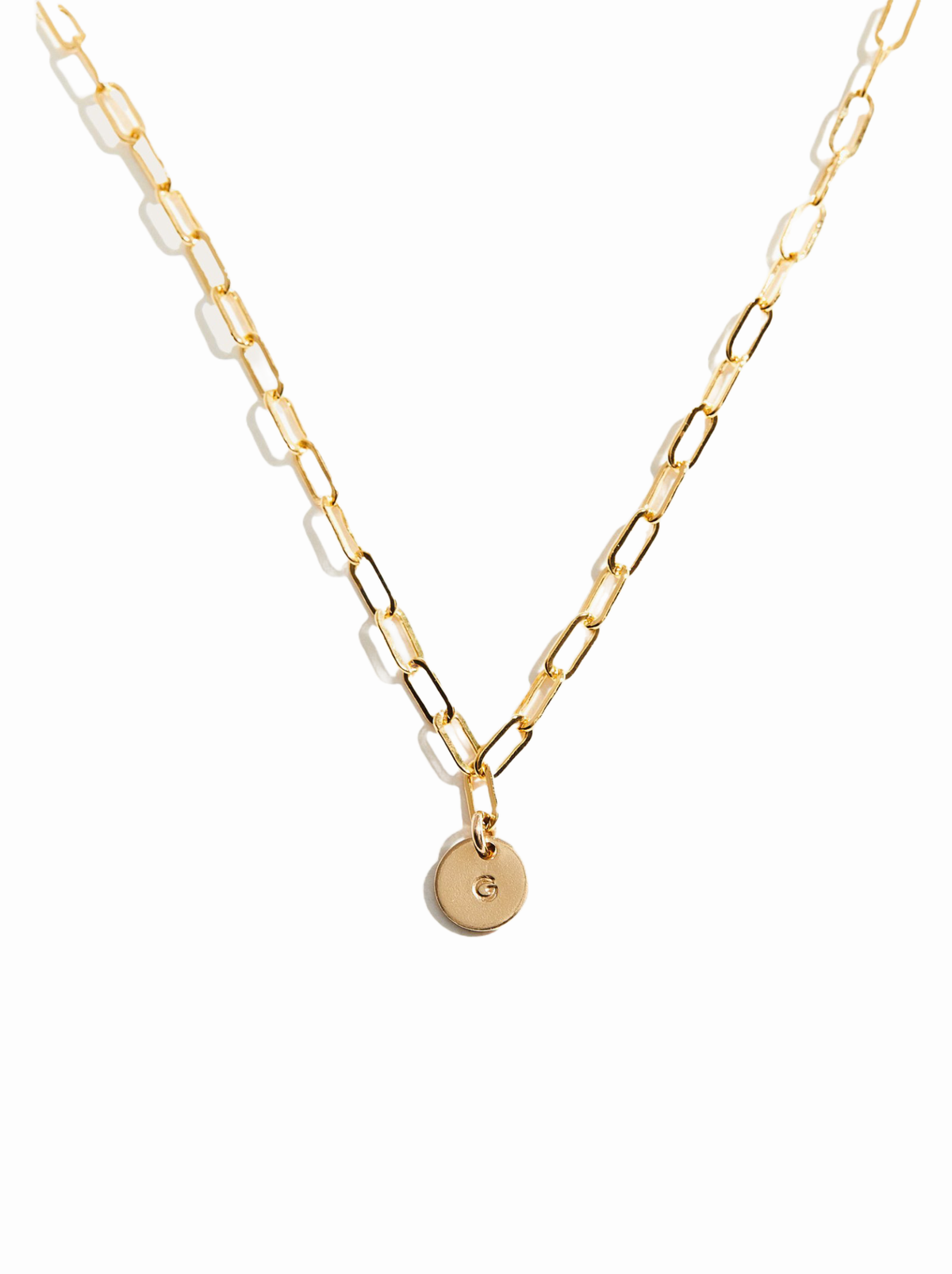 A gold paperclip chain necklace with a small round gold disc pendant featuring a stamped letter 'G', set against a plain white background.