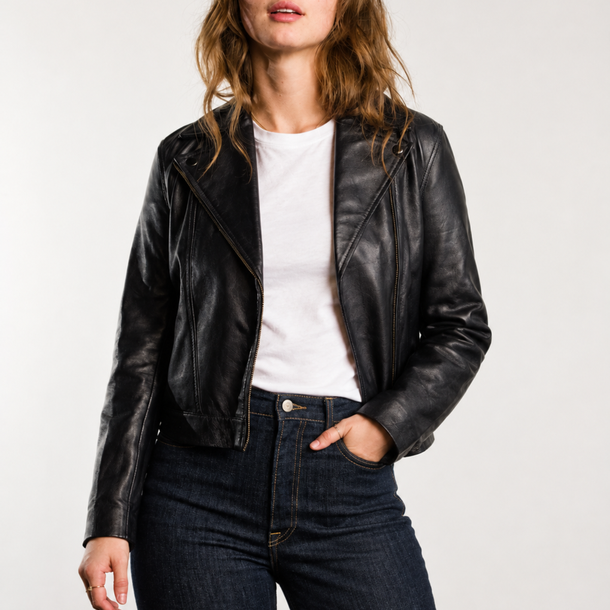 A woman wearing a black leather jacket, white tee, and jeans with one hand in her pocket against a white background.