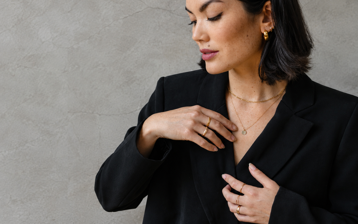 A woman in an oversized black blazer wearing layered jewelry styles from multiple earrings, necklaces and rings against a grey wall.