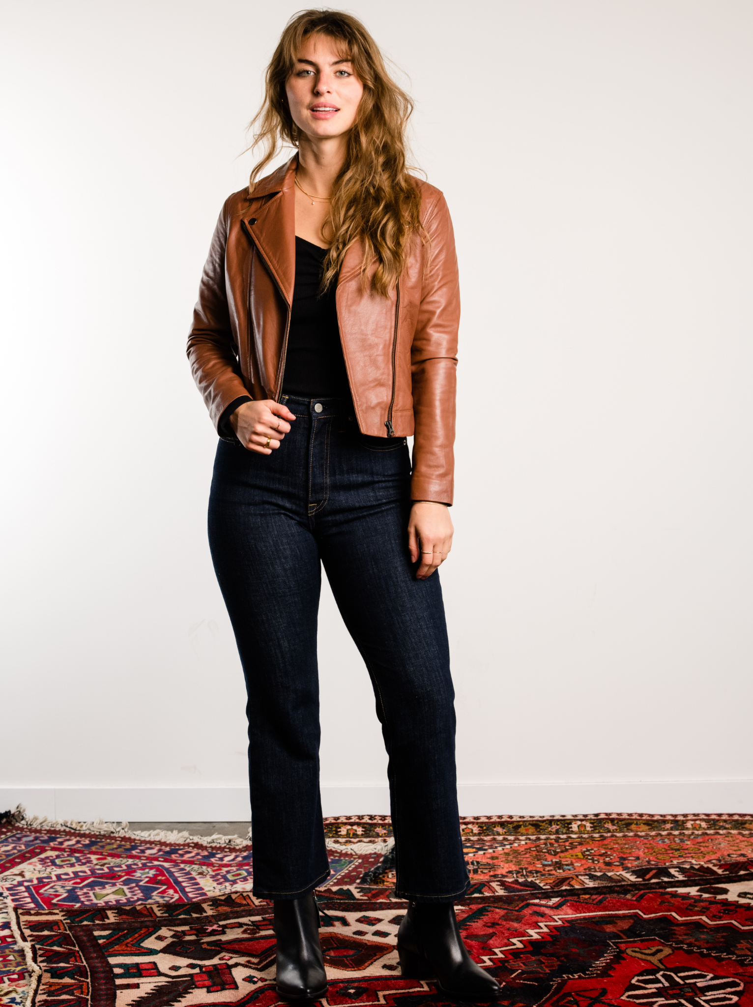 A person standing in front of a white background wearing a Whiskey Maha Leather Jacket, black top, black jeans, and black boots.
