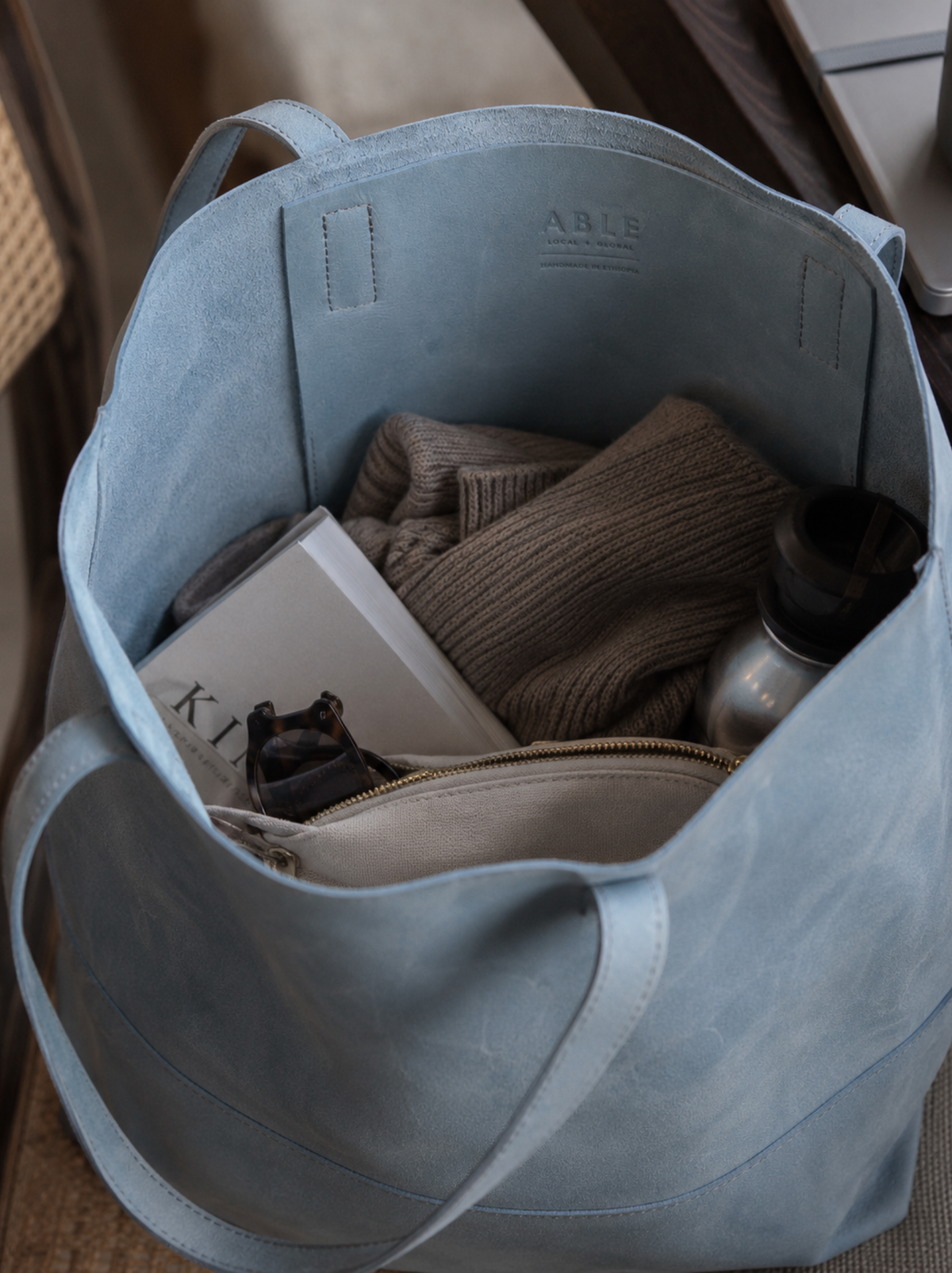 A light blue leather tote bag shown open, filled with a sweater, a book, a small pouch, and a water bottle. The brand name 'ABLE' is embossed on the inner slip pocket. #Color_Mist