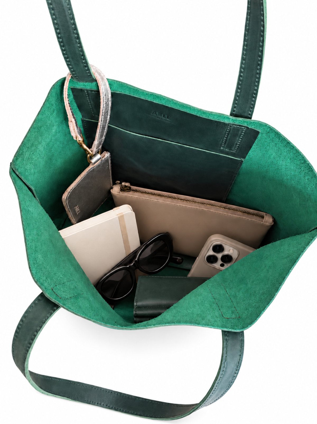 A green interior of a green leather tote with essential items inside, including a mini wristlet wrapped around the strap. #Color_Juniper