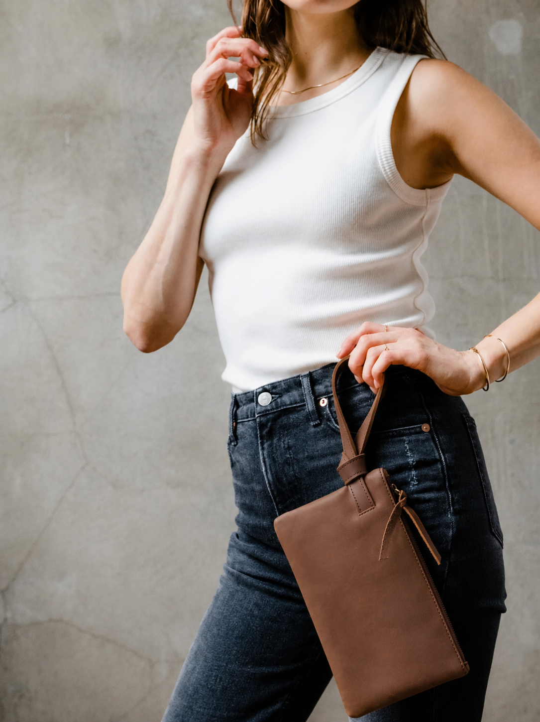 A woman wearing a white ribbed tank top and dark black jeans holds a brown leather wristlet by the handle against a textured gray concrete wall. #Color_Whiskey