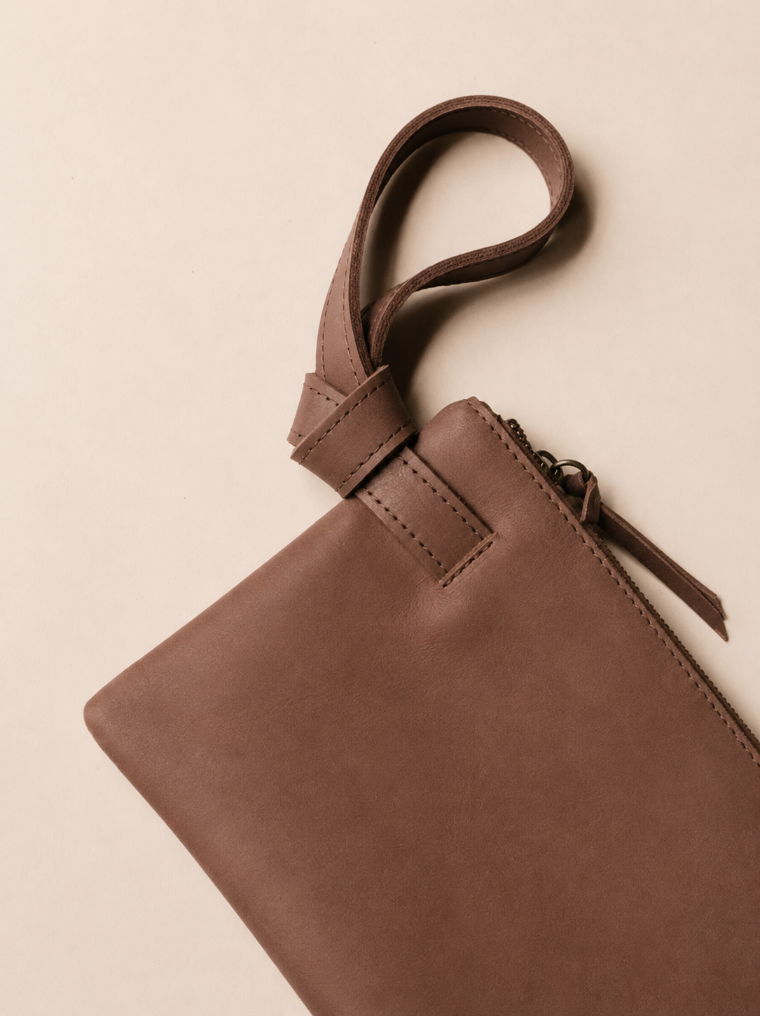 A close-up of a brown leather wristlet with a matching leather knotted loop handle, shown on a warm beige background. #Color_Whiskey
