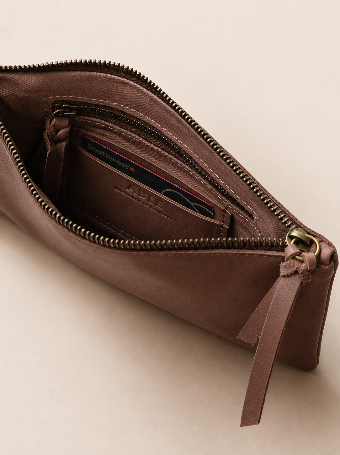 A close-up interior of a brown leather zippered wristlet with a matching leather pull tab, shown open to reveal an interior card slot containing a card. #Color_Whiskey