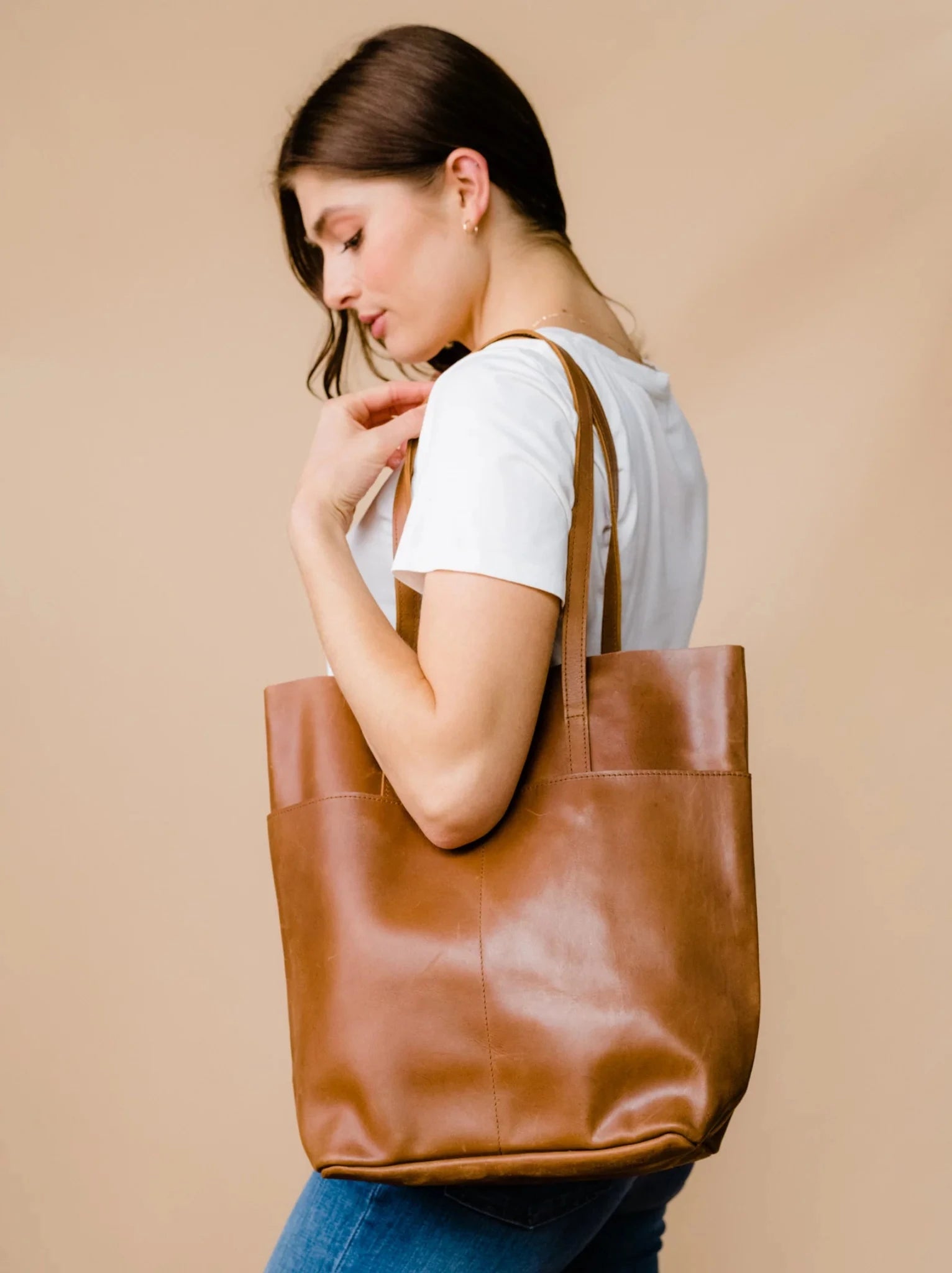 Woman carrying a brown leather tote bag on her shoulder in a tshirt and jeans against a neutral background. #Color_Whiskey