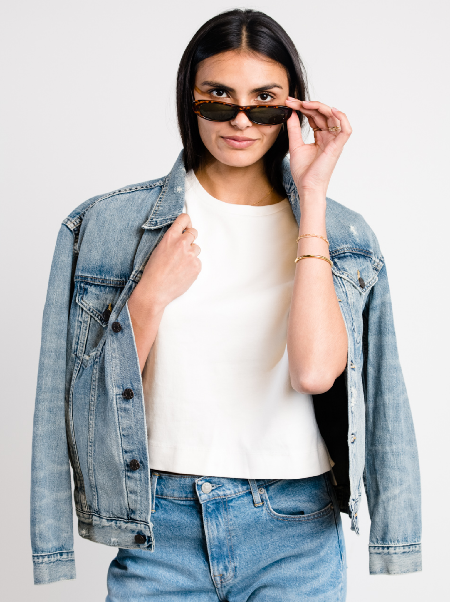 Woman wearing The Merly Jacket, white shirt, and sunglasses on a plain background